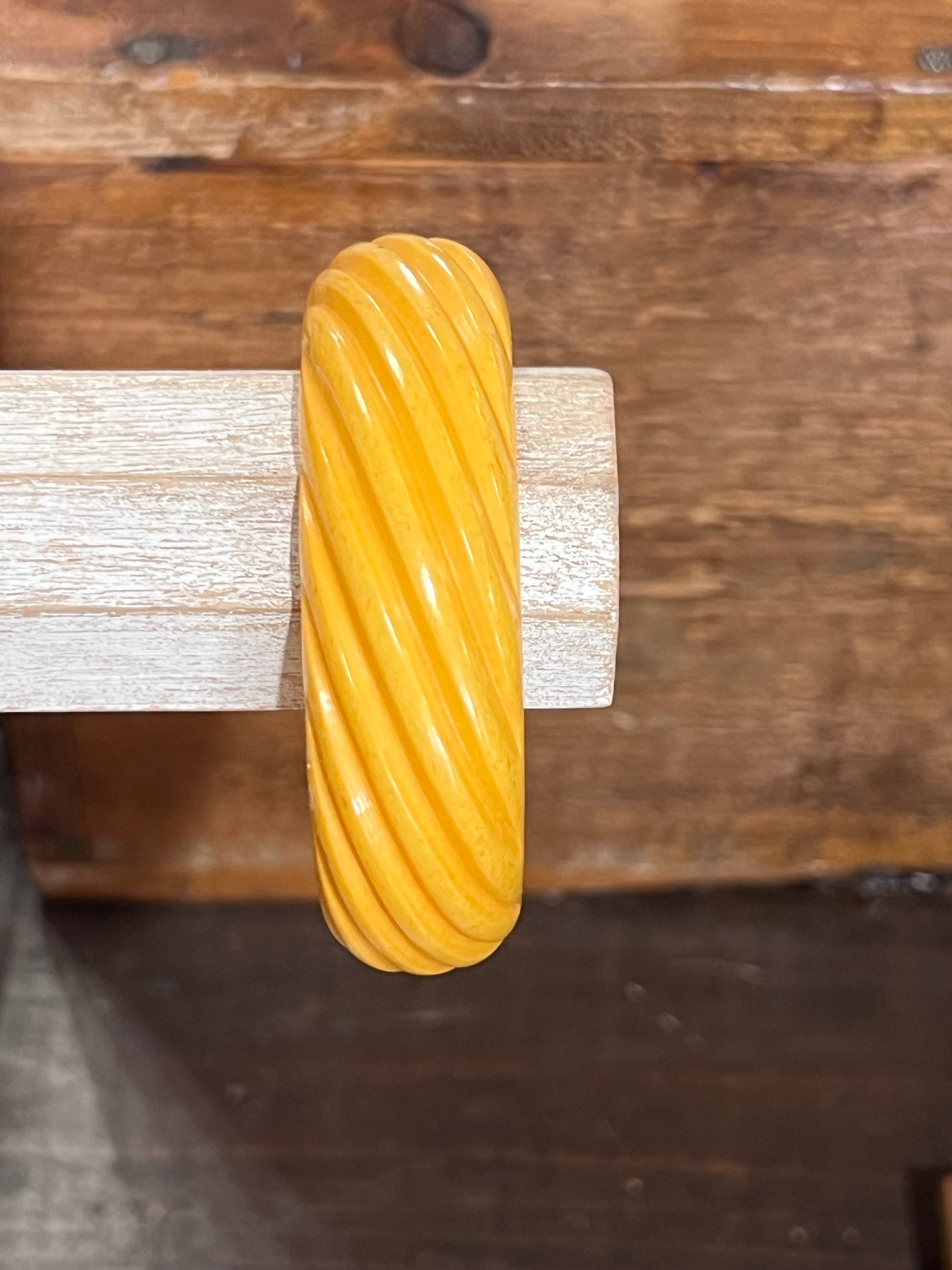 A thick, glossy yellow twisted bangle bracelet displayed on a light-colored wooden stand against a rustic dark wood background.