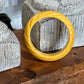 A twisted yellow bangle bracelet displayed on a rustic wooden surface in front of textured gray stone props.