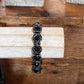 A black beaded stretch bracelet with faceted geometric beads, displayed on a light-colored wooden block over a rustic wooden surface.