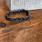 A black beaded stretch bracelet with round faceted beads, resting on a rustic wooden surface in front of a textured gray block.