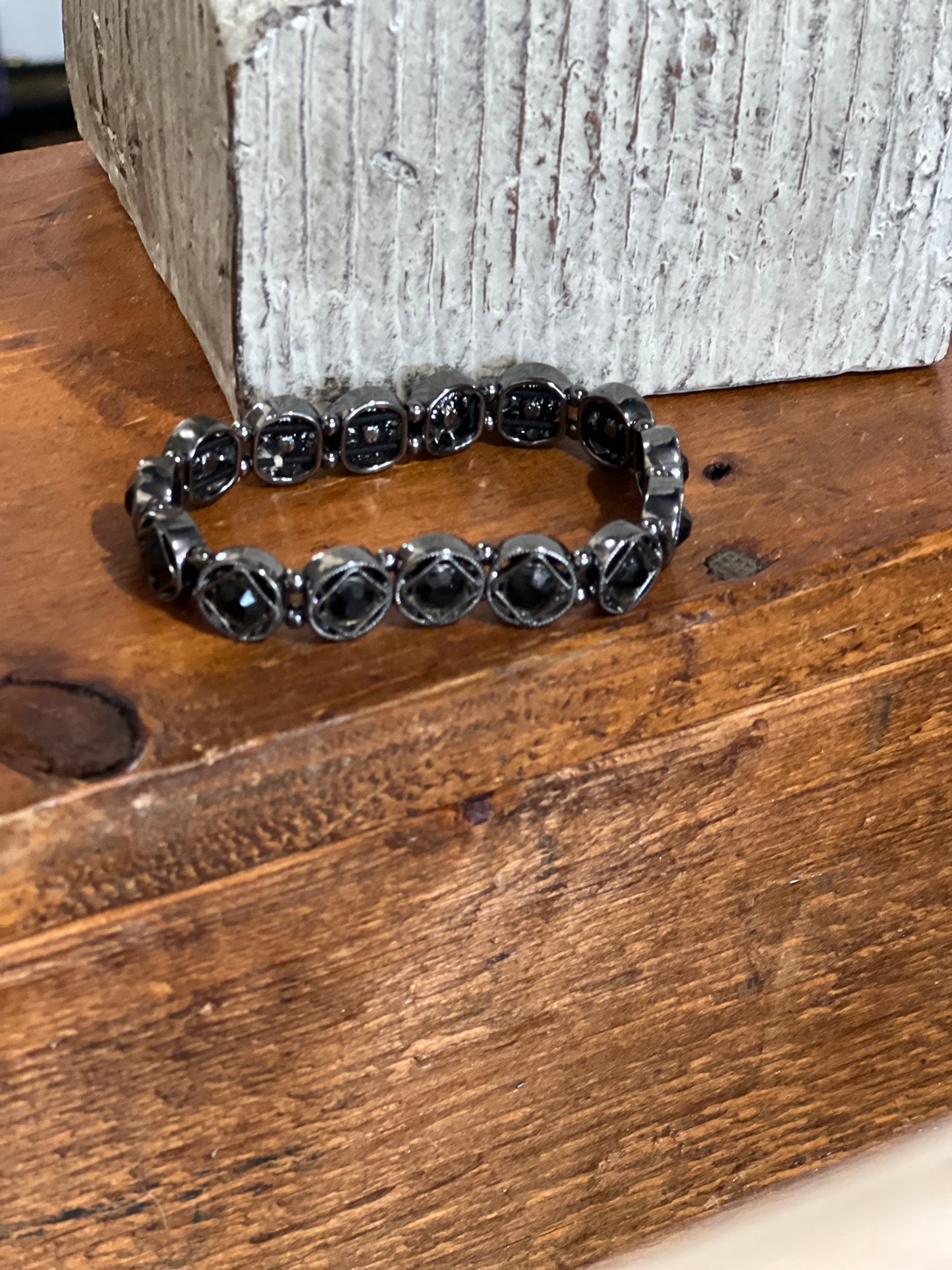 A black beaded stretch bracelet with round faceted beads, resting on a rustic wooden surface in front of a textured gray block.