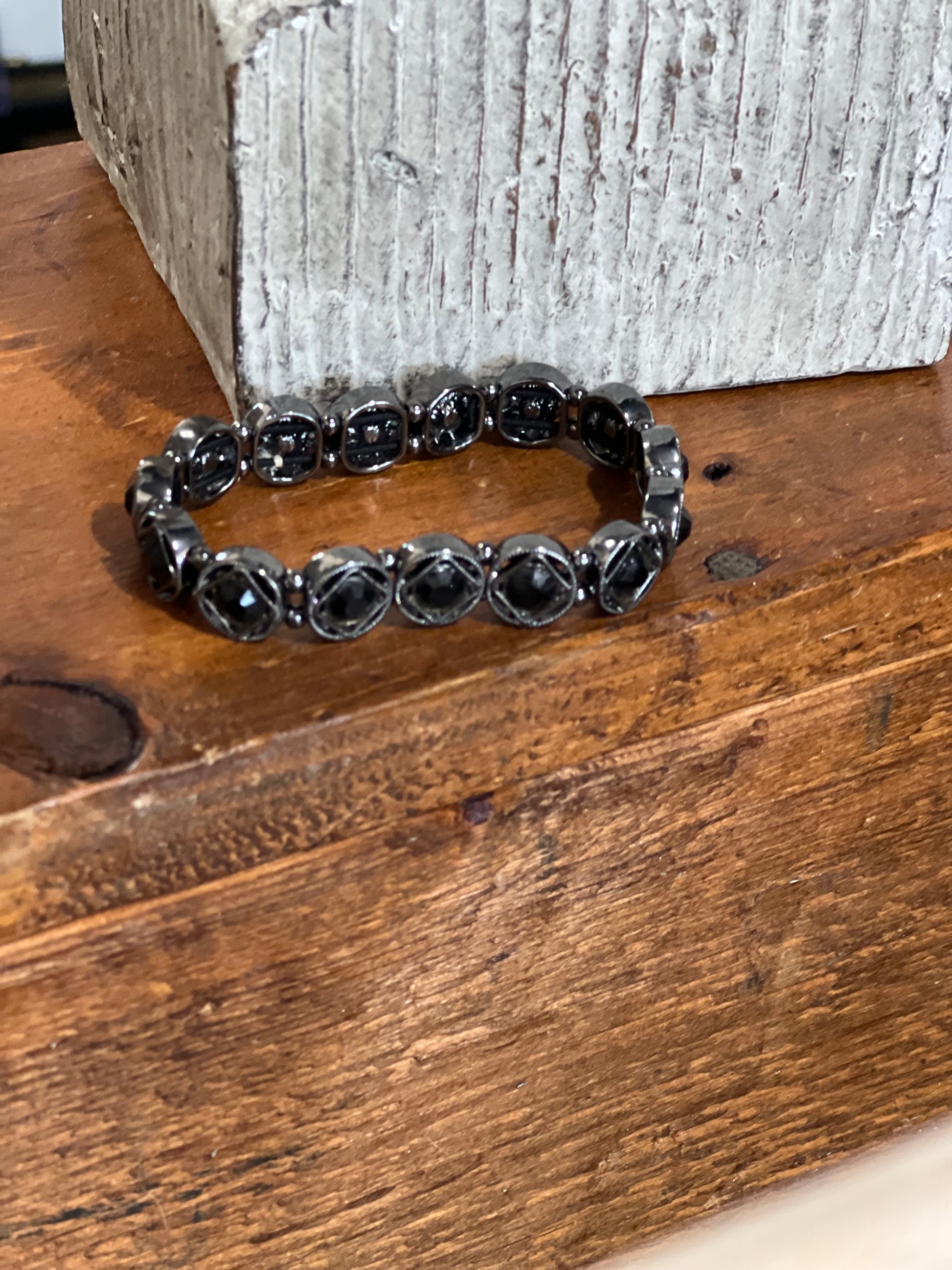 A black beaded stretch bracelet with round faceted beads, resting on a rustic wooden surface in front of a textured gray block.
