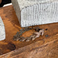 A bronze-toned leaf wreath bracelet resting on a rustic wooden table next to a textured gray stone block.