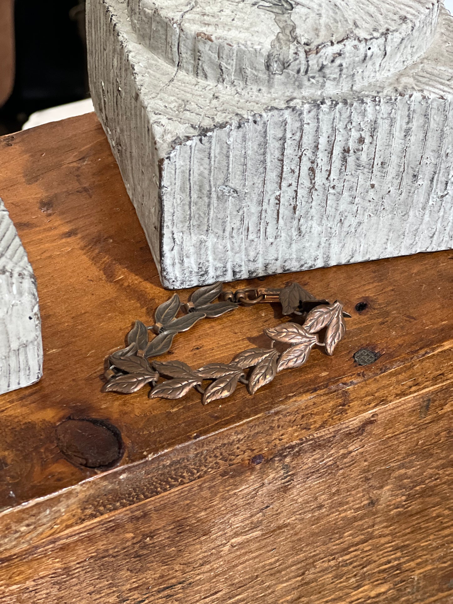 A bronze-toned leaf wreath bracelet resting on a rustic wooden table next to a textured gray stone block.