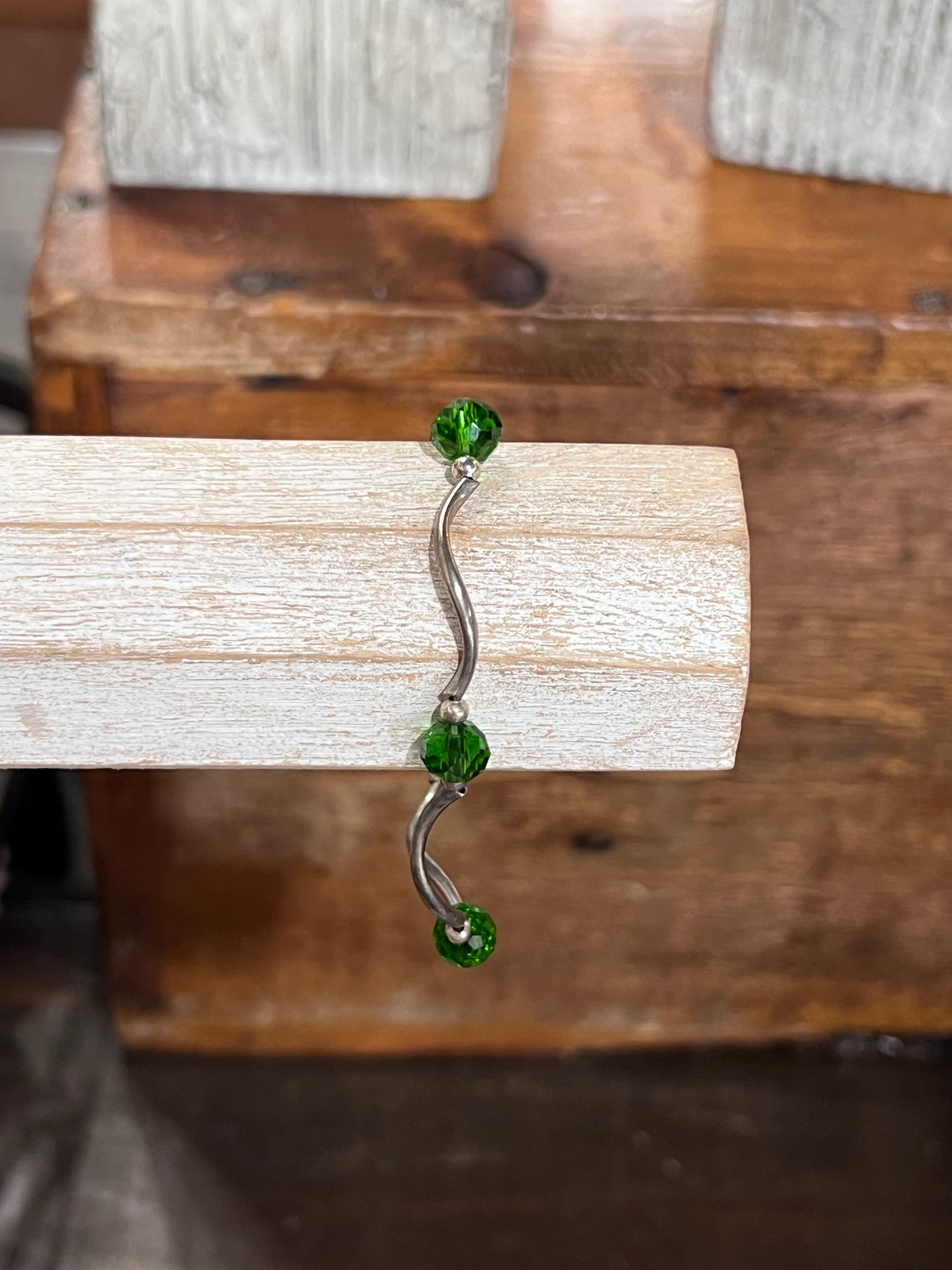 A silver-toned curved barbell nose ring with three faceted green gemstones, displayed on a light-colored wooden block against a rustic wooden background.