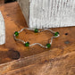 A silver wavy bracelet with five green faceted beads, displayed on a rustic wooden surface next to a textured gray block.