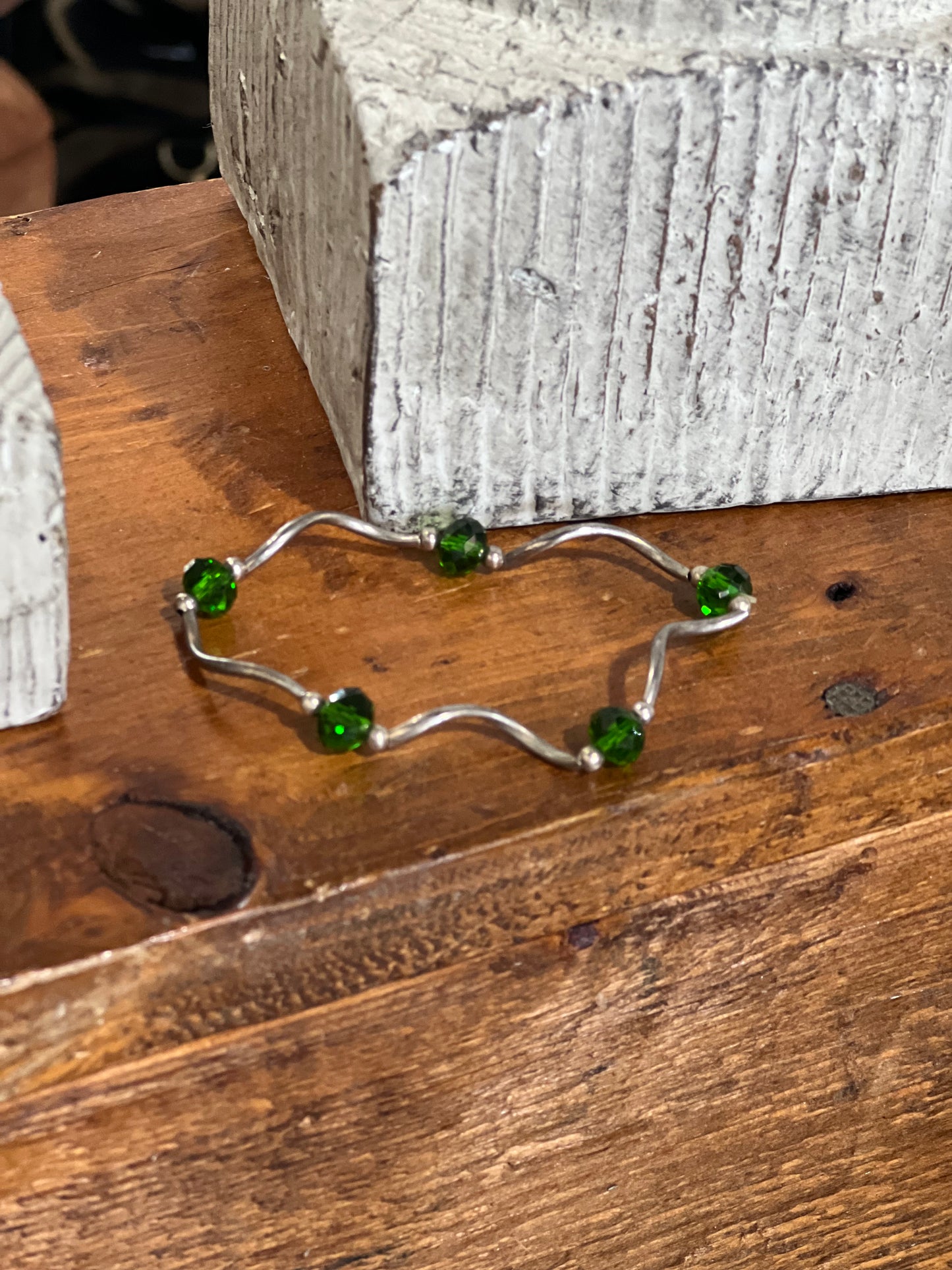 A silver wavy bracelet with five green faceted beads, displayed on a rustic wooden surface next to a textured gray block.