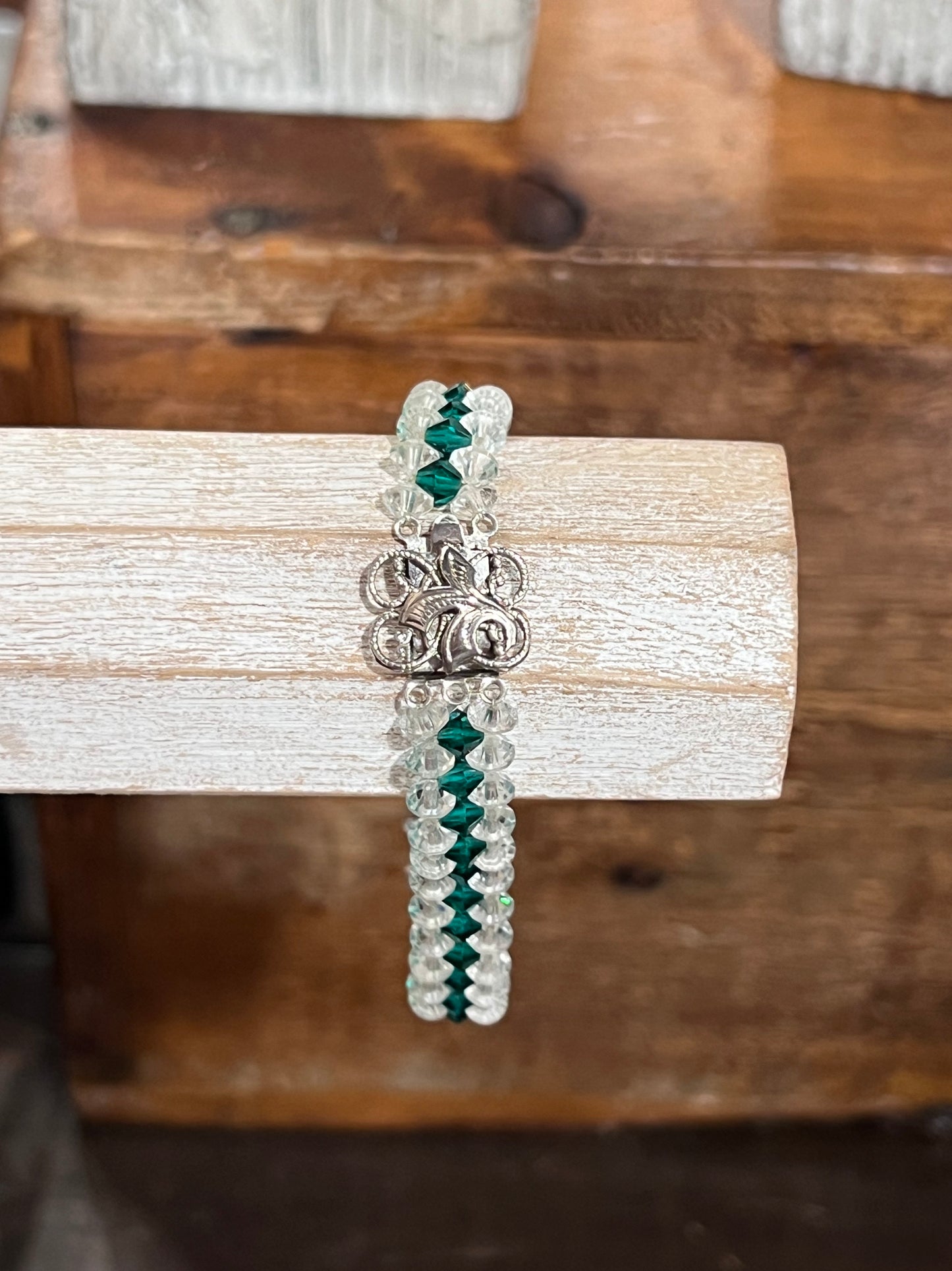 A beaded bracelet with silver-toned beads, dark green faceted beads, and a silver-toned floral clasp, displayed on a light wood block over a rustic wooden background.