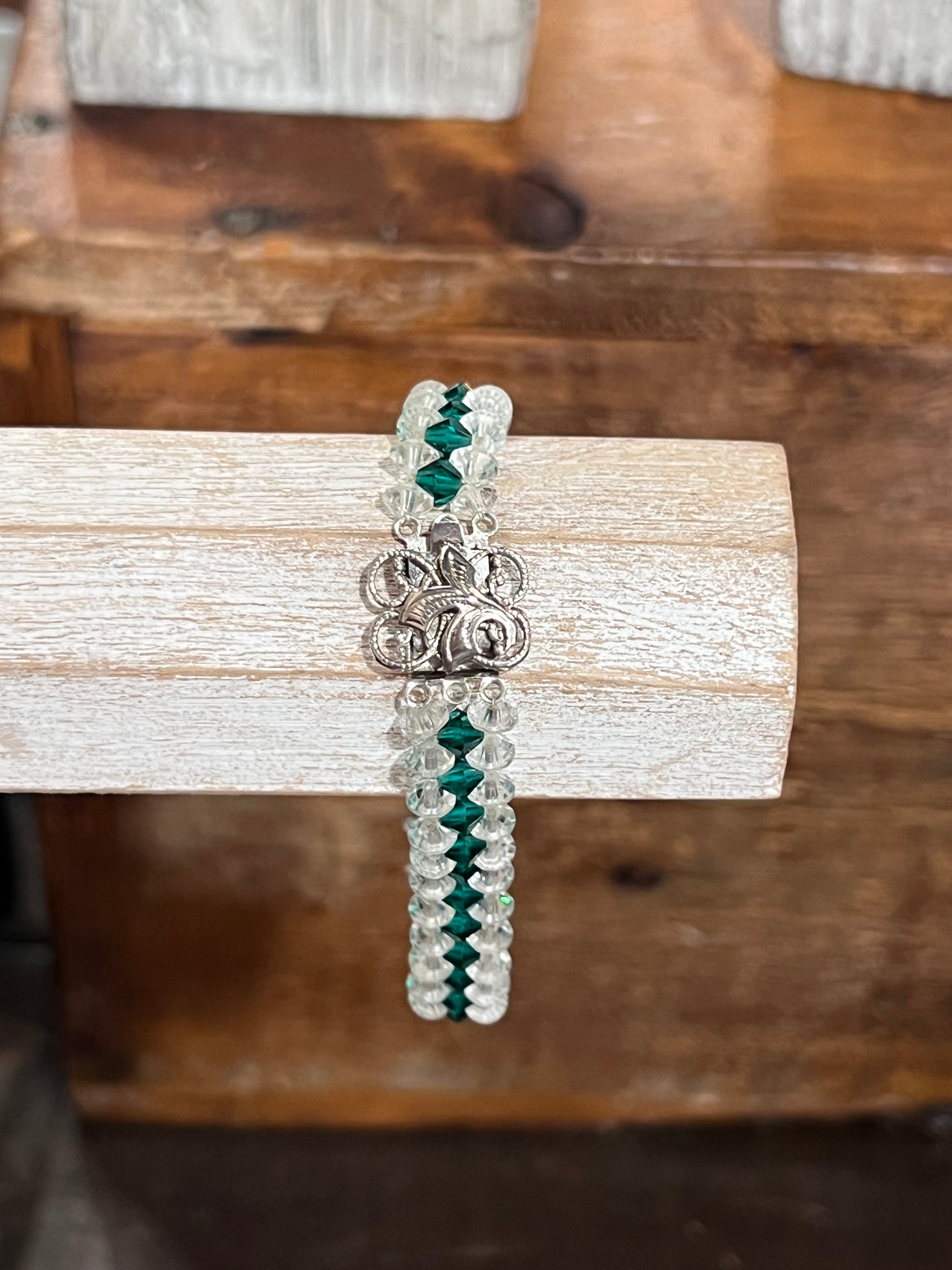 A beaded bracelet with silver-toned beads, dark green faceted beads, and a silver-toned floral clasp, displayed on a light wood block over a rustic wooden background.