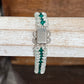 A beaded bracelet with clear faceted beads, green crystal accents, and a silver-toned metal clasp, displayed on a rustic white wooden stand.