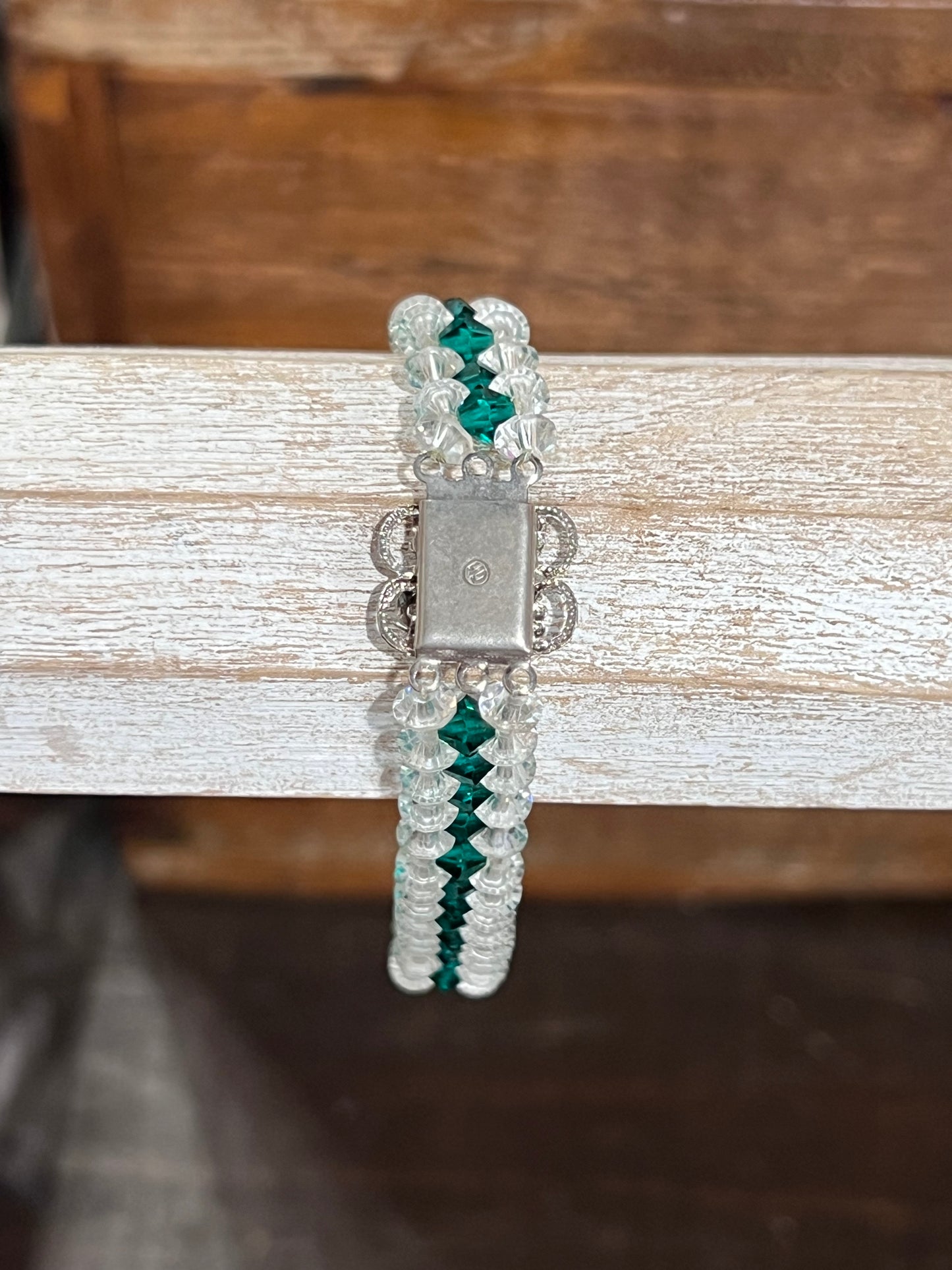 A beaded bracelet with clear faceted beads, green crystal accents, and a silver-toned metal clasp, displayed on a rustic white wooden stand.