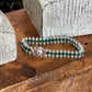 A double-strand beaded bracelet with green faceted beads, clear crystal-like beads, and a silver-toned floral clasp, displayed on a rustic wooden surface.