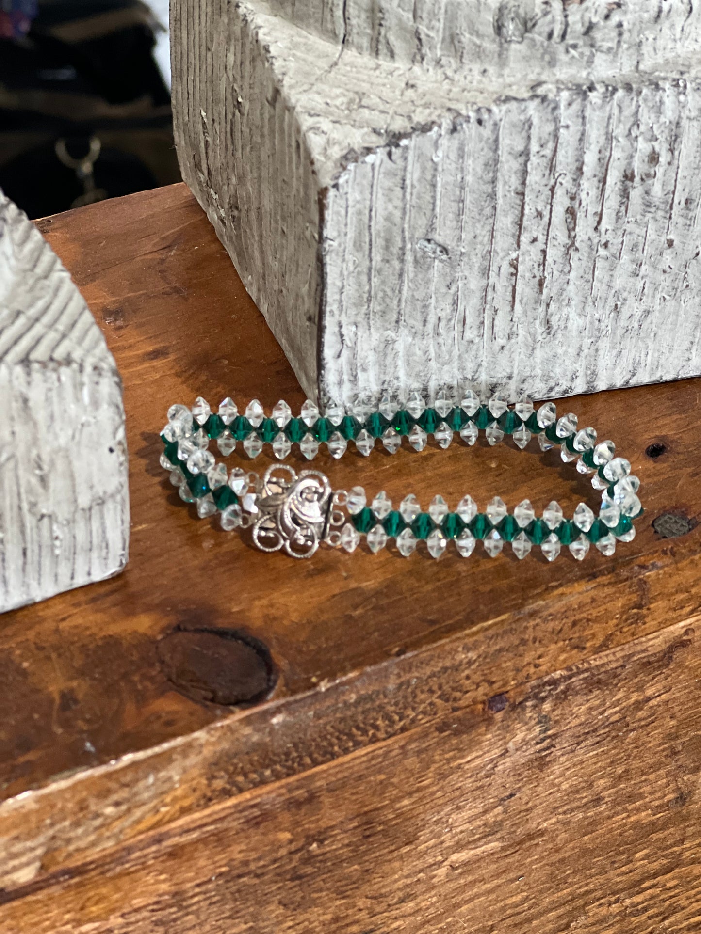 A double-strand beaded bracelet with green faceted beads, clear crystal-like beads, and a silver-toned floral clasp, displayed on a rustic wooden surface.