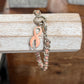 A silver-toned chain bracelet with a pink enamel ribbon charm, displayed hanging from a rustic white wooden block over a dark wood surface.