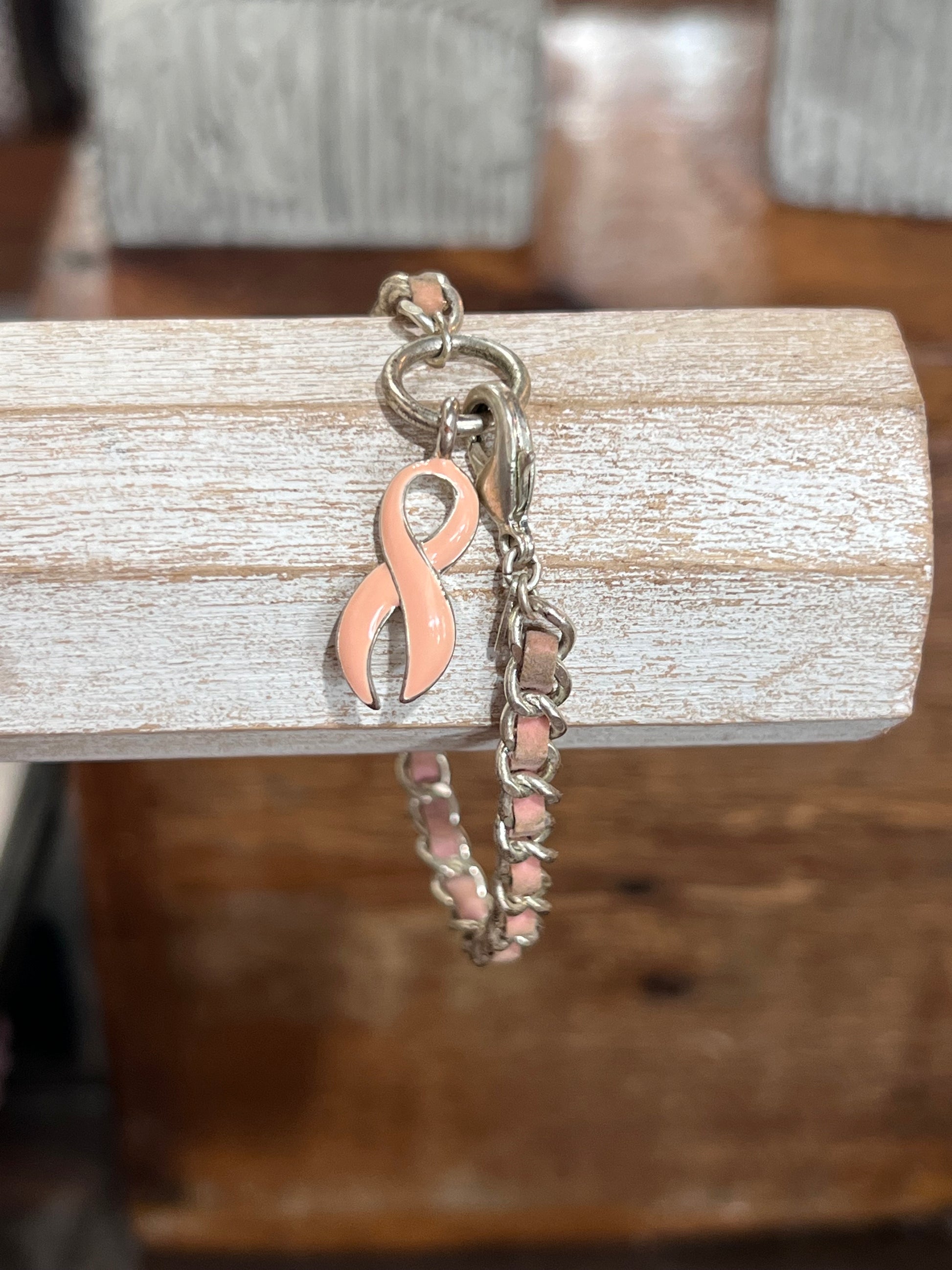 A silver-toned chain bracelet with a pink enamel ribbon charm, displayed hanging from a rustic white wooden block over a dark wood surface.