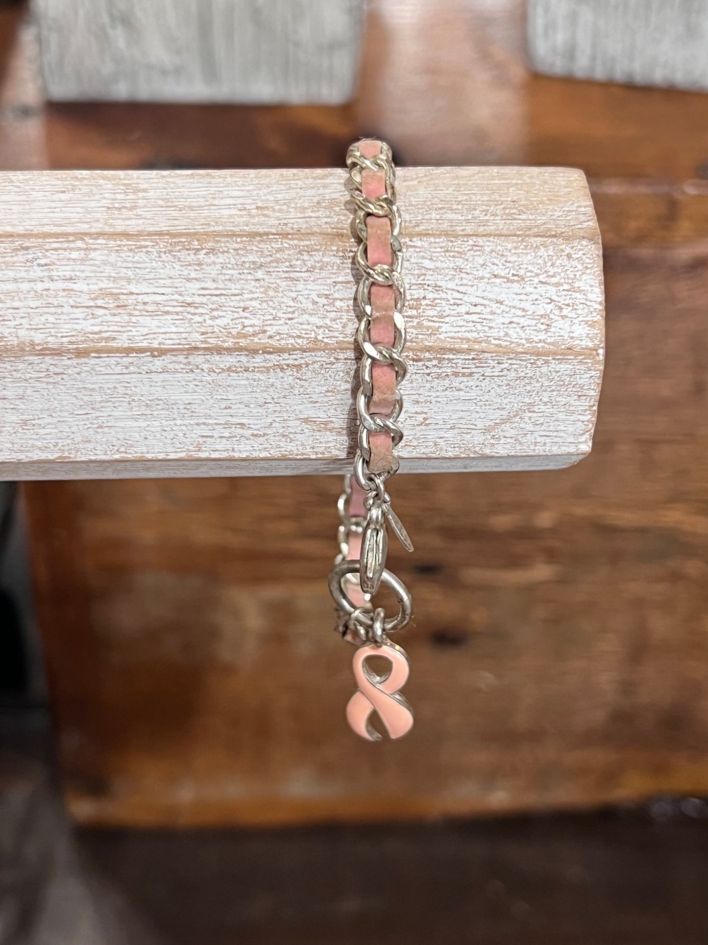 A silver-toned chain bracelet with a peach-pink link accent and a dangling pink awareness ribbon charm, displayed on a rustic wooden block.