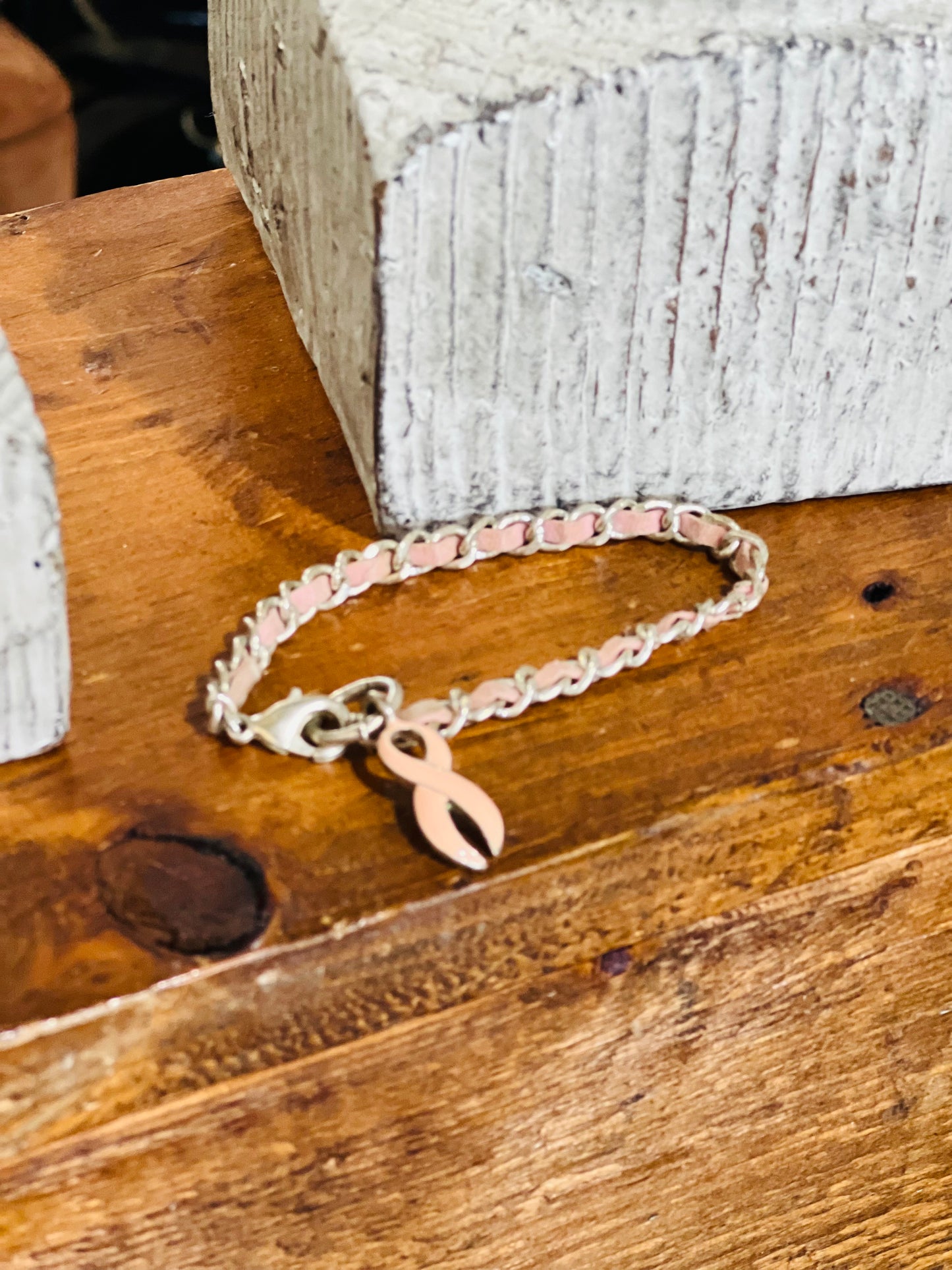 A silver-toned chain bracelet with a pink awareness ribbon charm, displayed on a rustic wooden surface next to a white textured block.