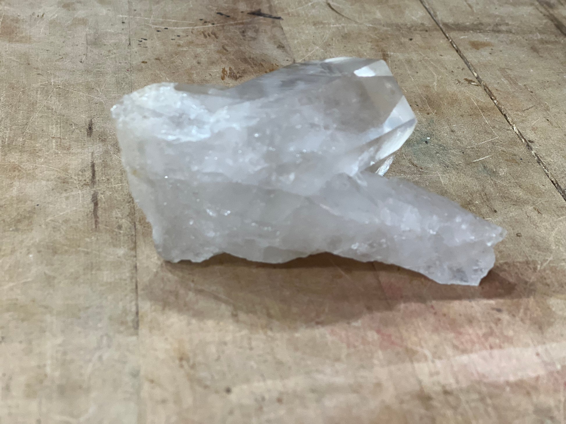 A rough, unpolished crystal cluster with natural faceted edges, resting on a textured wooden surface.