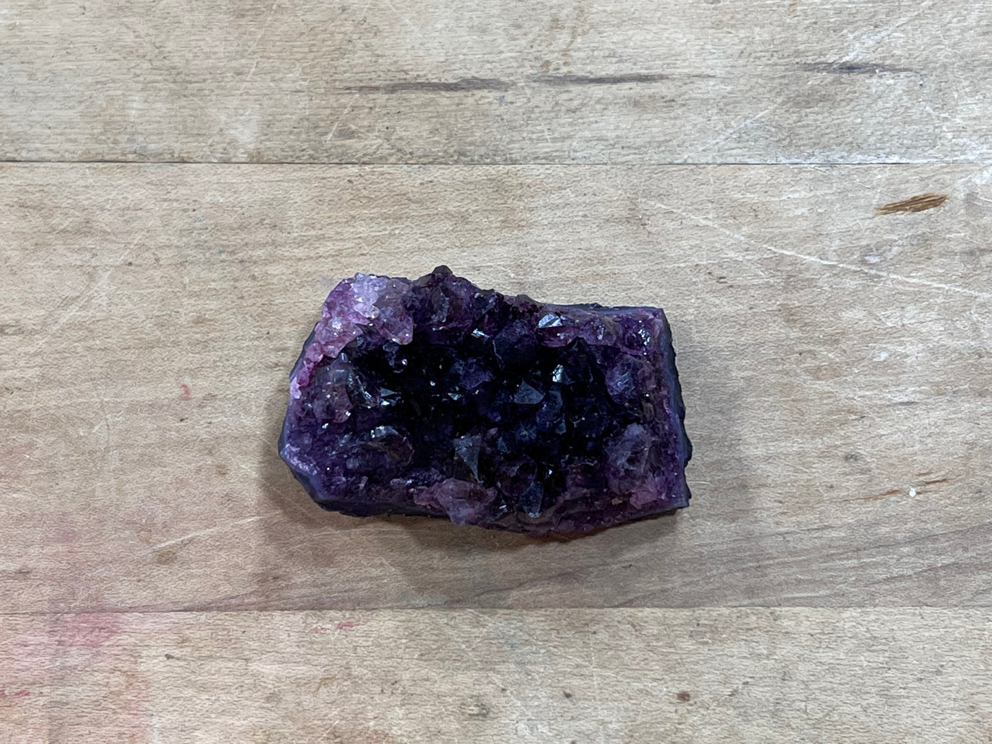 A raw amethyst crystal cluster with a dark purple cavity and faceted purple crystal points, resting on a light brown wooden surface.