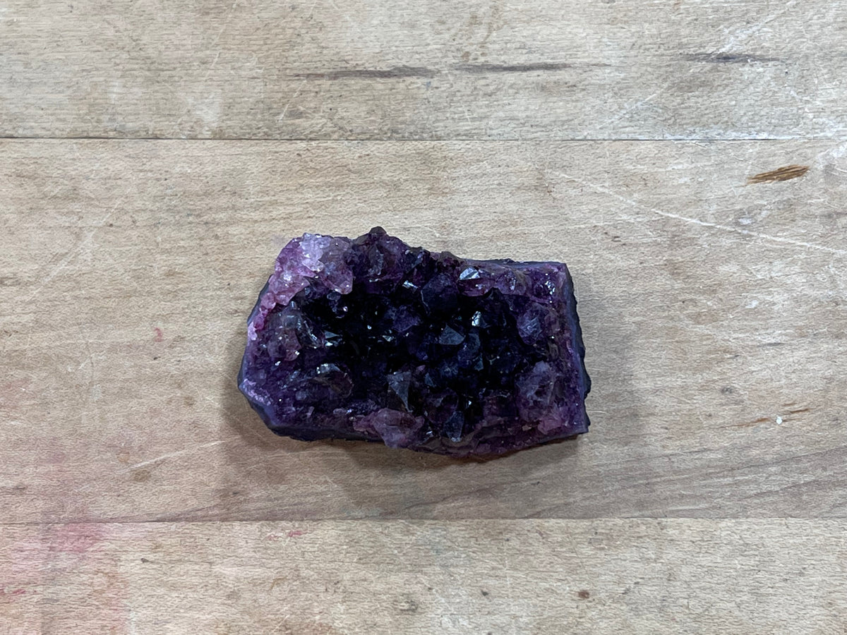 A raw amethyst crystal cluster with a dark purple cavity and faceted purple crystal points, resting on a light brown wooden surface.