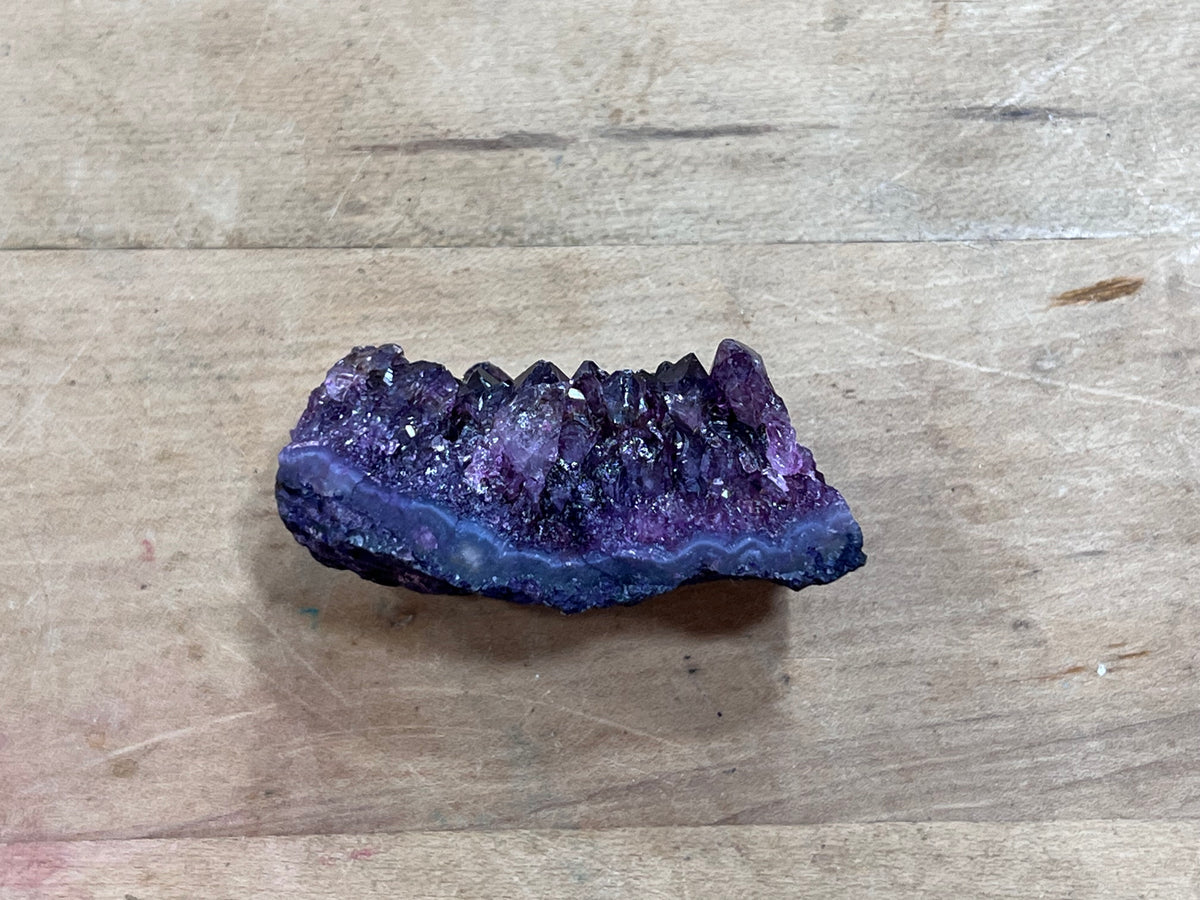 A raw purple crystal cluster geode section with dark blue and violet agate bands, resting on a light brown wooden surface.