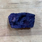 A raw, rough purple mineral specimen with a dark, textured surface, resting on a light-colored wooden surface.