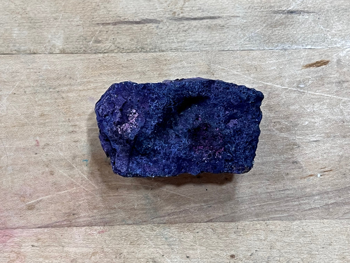 A raw, rough purple mineral specimen with a dark, textured surface, resting on a light-colored wooden surface.