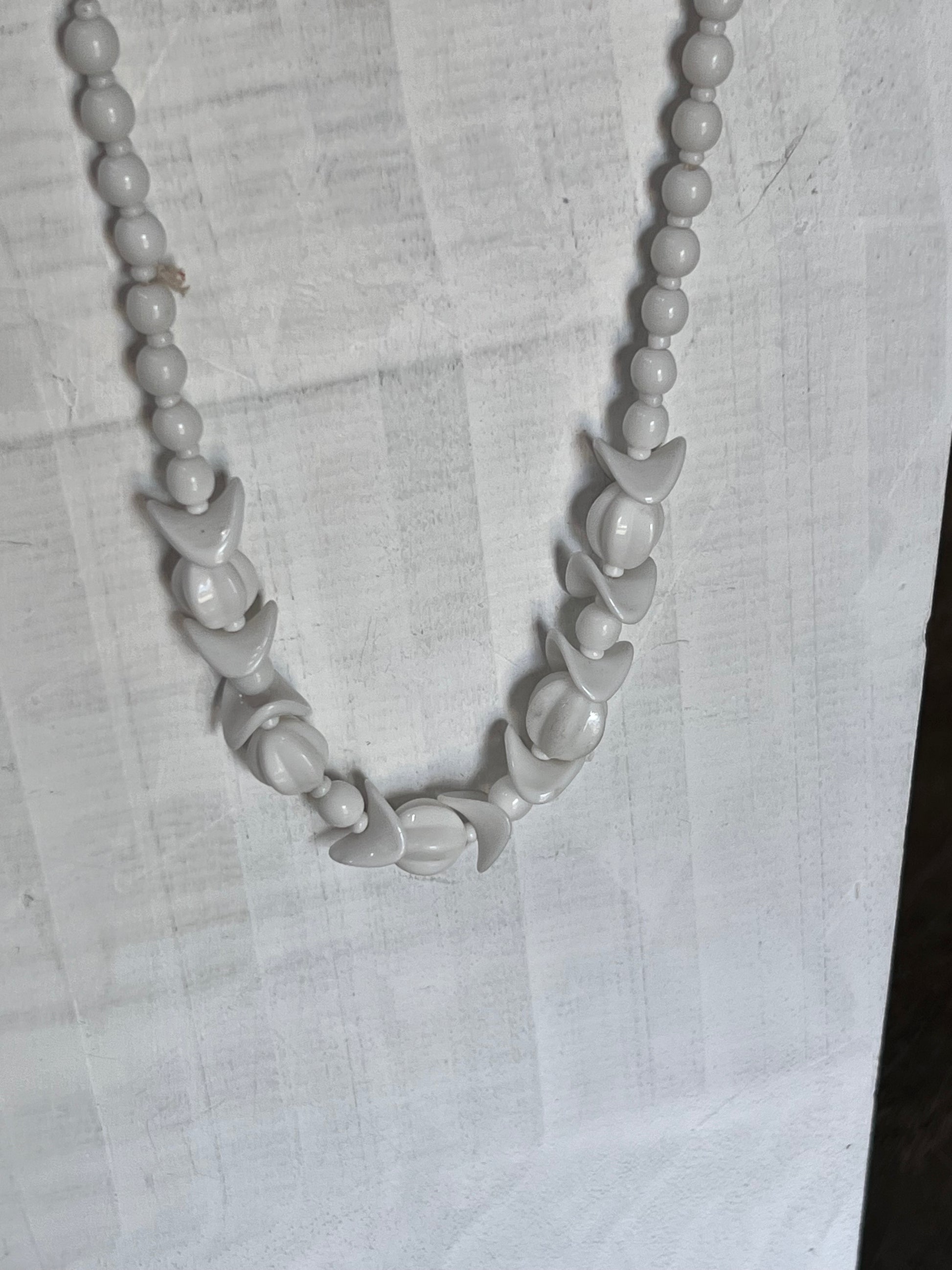 A white beaded necklace with alternating smooth round beads and carved shell-like teardrop beads, displayed on a wrinkled white fabric background.