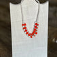 A beaded necklace with white round beads and bright red-orange bead clusters, displayed on a white fabric jewelry bust against a blurred brown background.