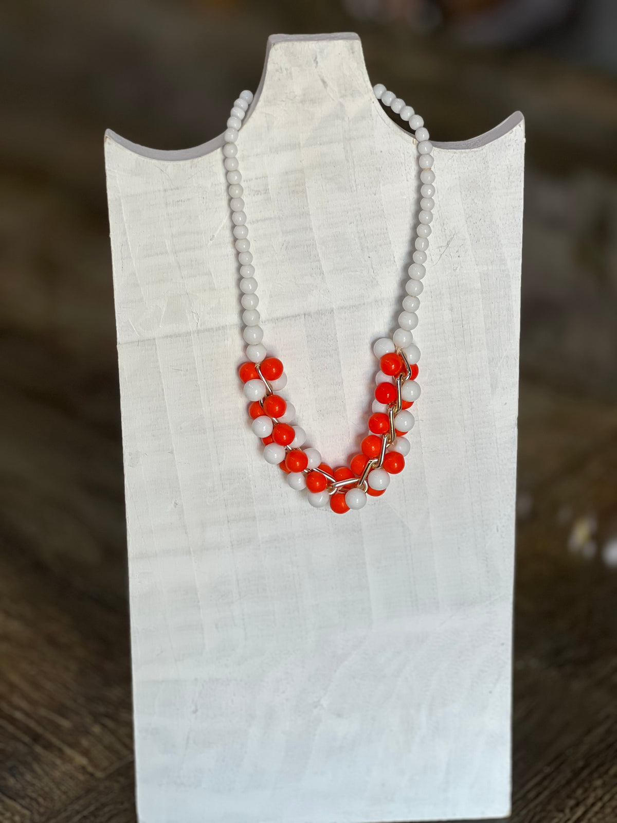A beaded necklace with white round beads and bright red-orange bead clusters, displayed on a white fabric jewelry bust against a blurred brown background.