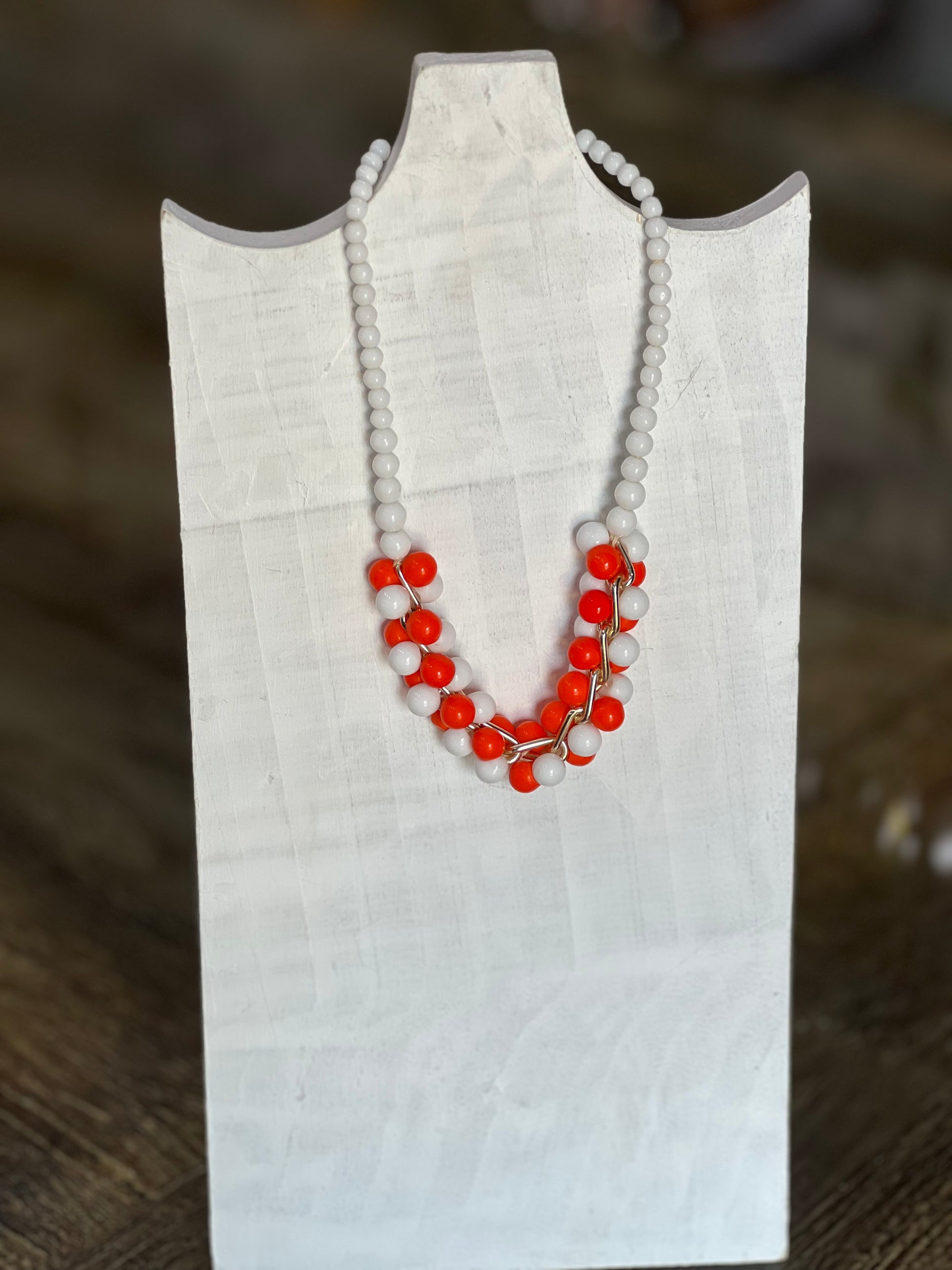 A beaded necklace with white round beads and bright red-orange bead clusters, displayed on a white fabric jewelry bust against a blurred brown background.