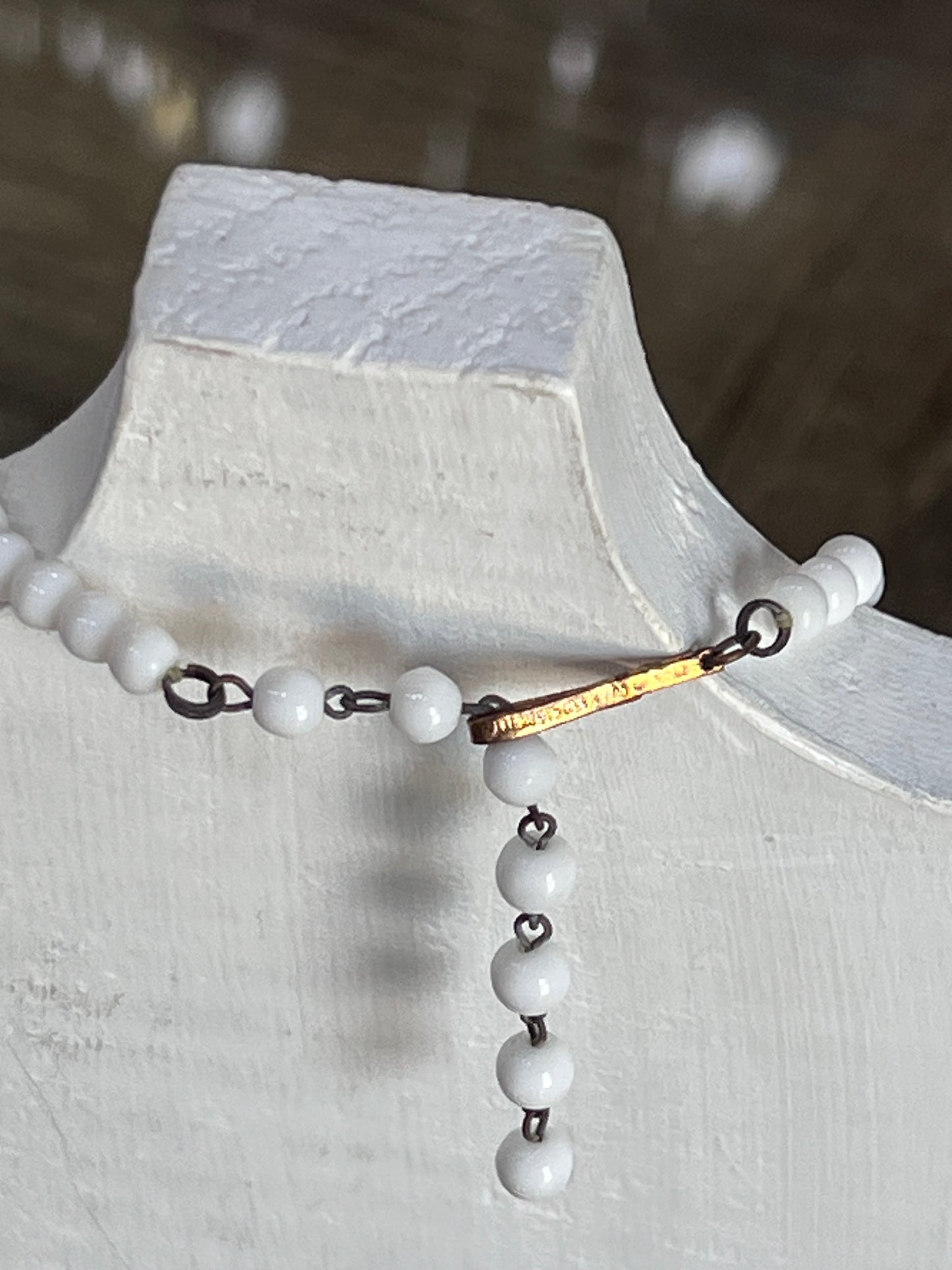 A beaded necklace with white round beads, dark metal links, and a small gold-toned bar detail, displayed on a white jewelry bust.