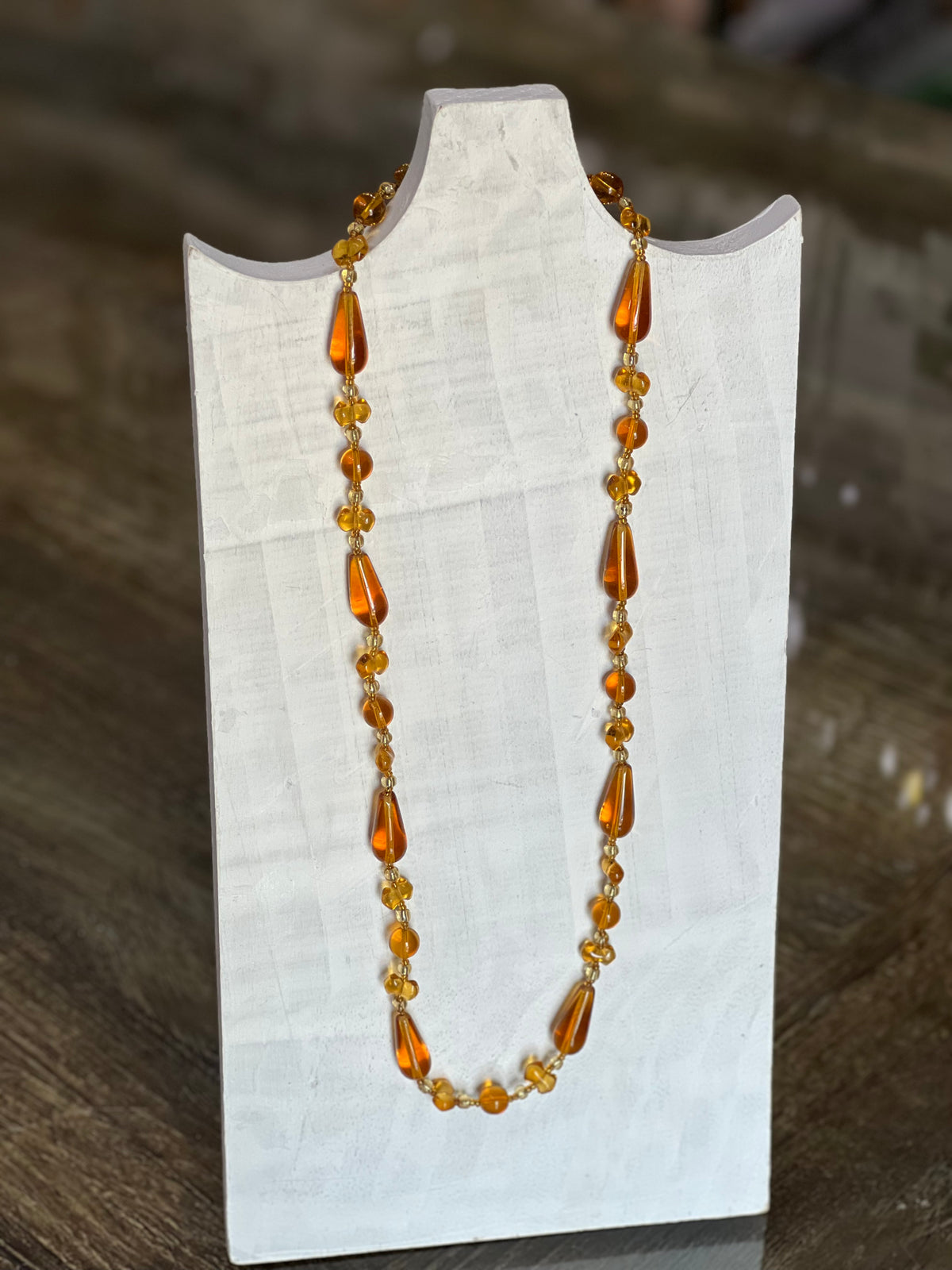 An amber glass bead necklace with teardrop-shaped beads and small round beads, displayed on a white fabric-covered jewelry stand against a dark wooden background.