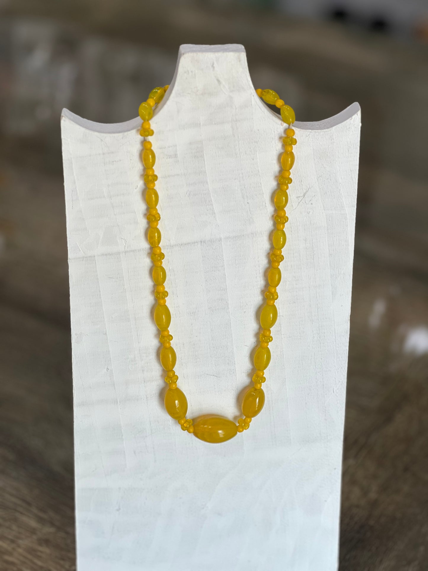A yellow beaded necklace displayed on a white fabric-covered jewelry stand against a blurred brown wooden background.