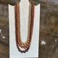 Three-strand pearl necklace with warm gradient beads transitioning from light champagne to medium brown and dark chocolate, displayed on a white stand over a wooden surface.
