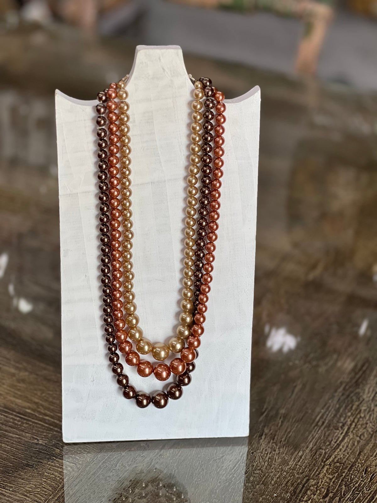 Three-strand pearl necklace with warm gradient beads transitioning from light champagne to medium brown and dark chocolate, displayed on a white stand over a wooden surface.