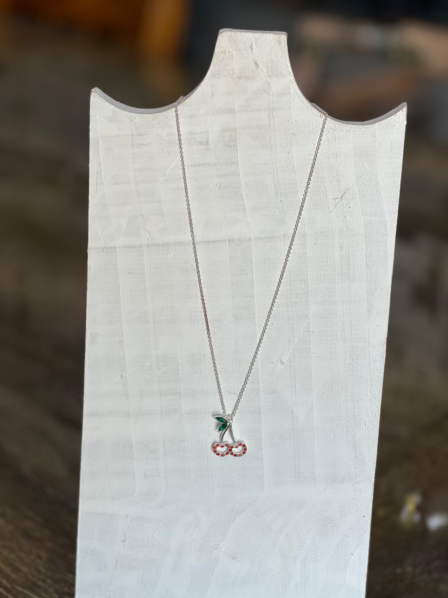 A silver necklace with a cherry pendant displayed on a white fabric jewelry stand against a blurred wooden background.
