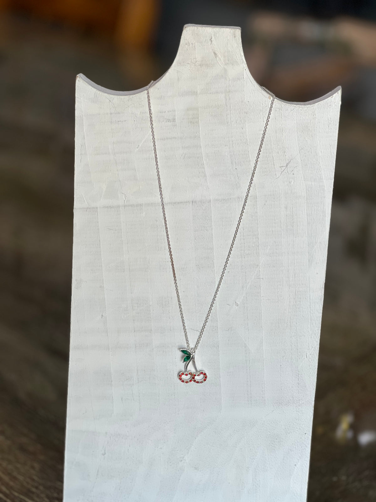 A silver necklace with a cherry pendant displayed on a white fabric jewelry stand against a blurred wooden background.