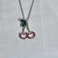 A silver-tone cherry pendant necklace with a green leaf and red rhinestone-studded cherries on a textured white background.