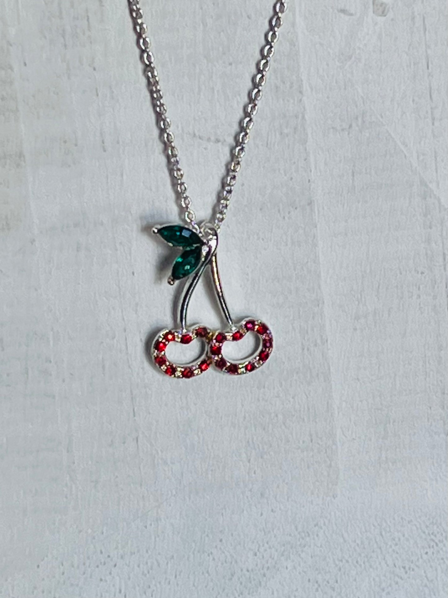 A silver-tone cherry pendant necklace with a green leaf and red rhinestone-studded cherries on a textured white background.