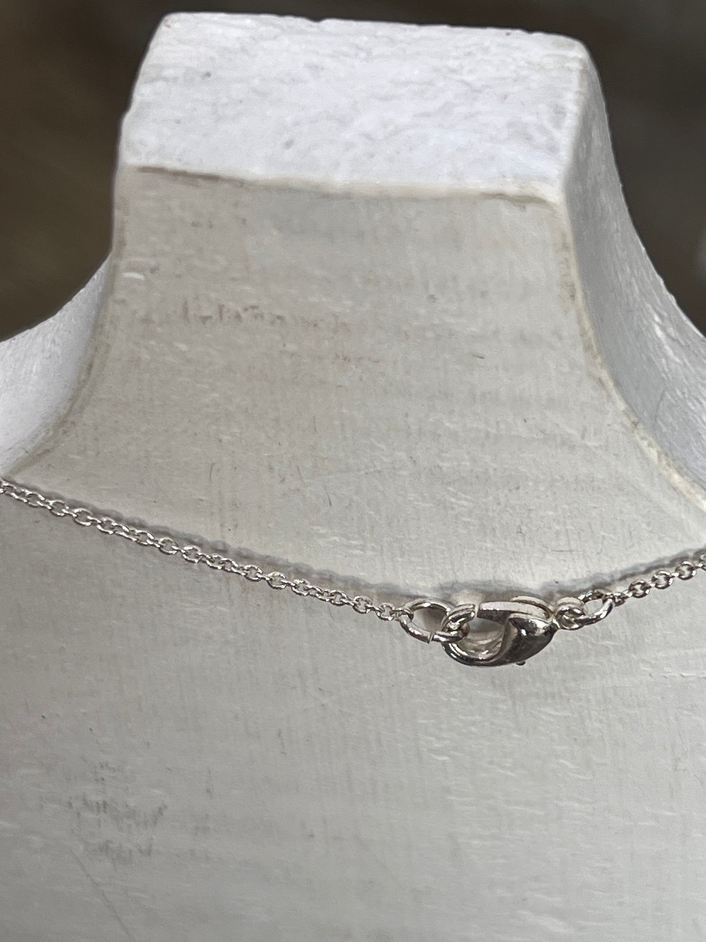 A delicate silver chain necklace with a small heart-shaped charm and clasp, displayed on a white textured mannequin bust.