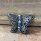 A butterfly-shaped filigree brooch with intricate dark metalwork, featuring blue faceted beads and small silver spacer beads down the center, displayed on a rustic wooden surface.