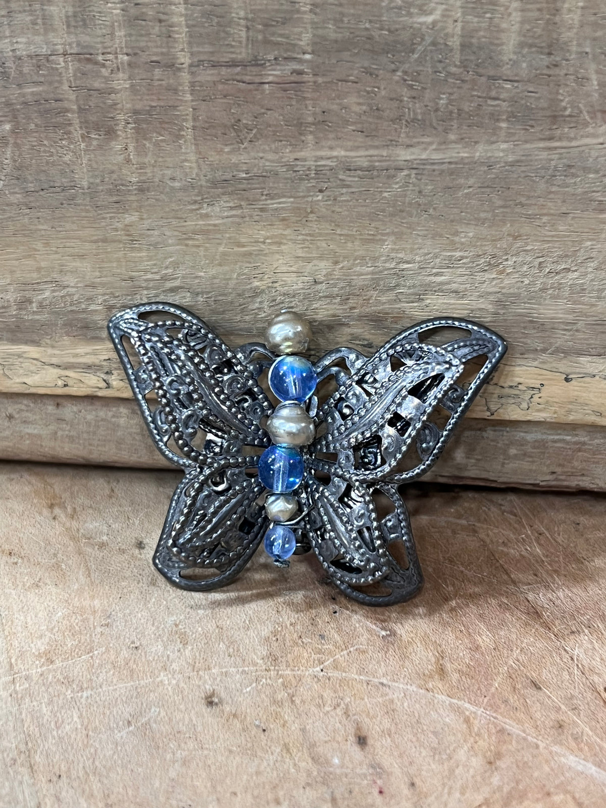 A butterfly-shaped filigree brooch with intricate dark metalwork, featuring blue faceted beads and small silver spacer beads down the center, displayed on a rustic wooden surface.