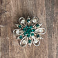 A vintage-style silver-tone brooch shaped like a flower, featuring a central cluster of green rhinestones surrounded by smaller clear and green stones, displayed on a rustic wooden surface.