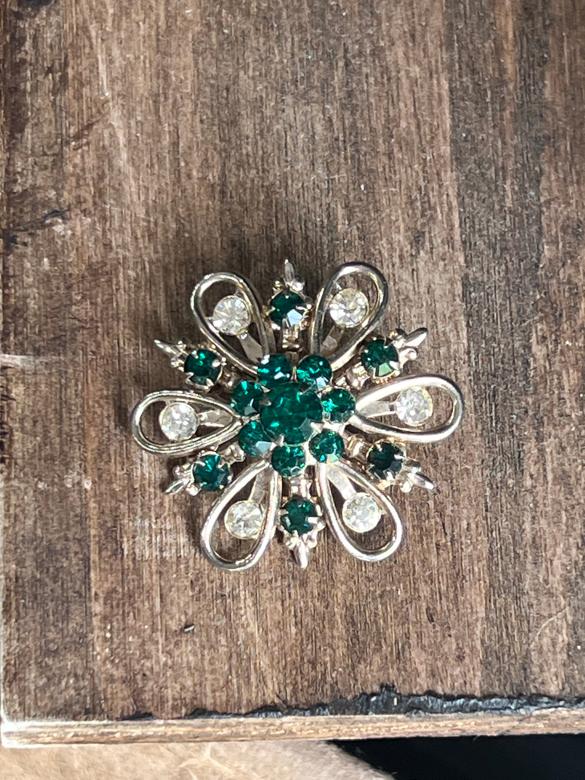 A vintage-style silver-tone brooch shaped like a flower, featuring a central cluster of green rhinestones surrounded by smaller clear and green stones, displayed on a rustic wooden surface.