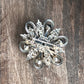 An ornate silver-toned metal brooch with a vintage floral or scroll design, displayed on a dark brown wooden surface.