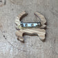 A small wooden charm or brooch with a natural wood grain finish and a silver metal safety pin clasp, shown on a light brown wooden surface.