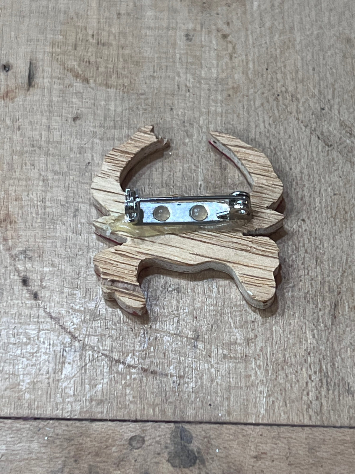 A small wooden charm or brooch with a natural wood grain finish and a silver metal safety pin clasp, shown on a light brown wooden surface.