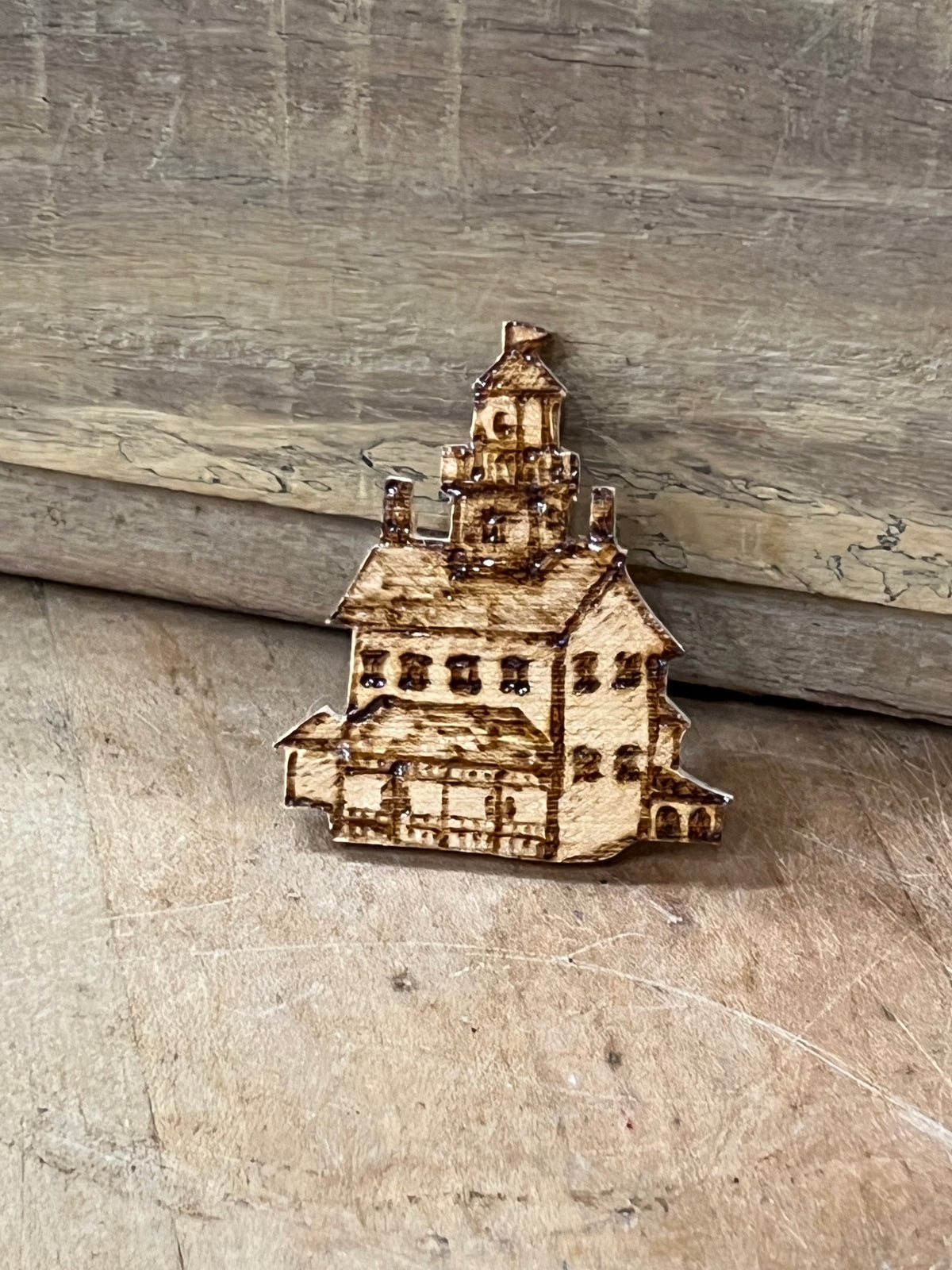 A small wooden house-shaped ornament or charm with laser-burned details, resting on a rustic wooden surface.