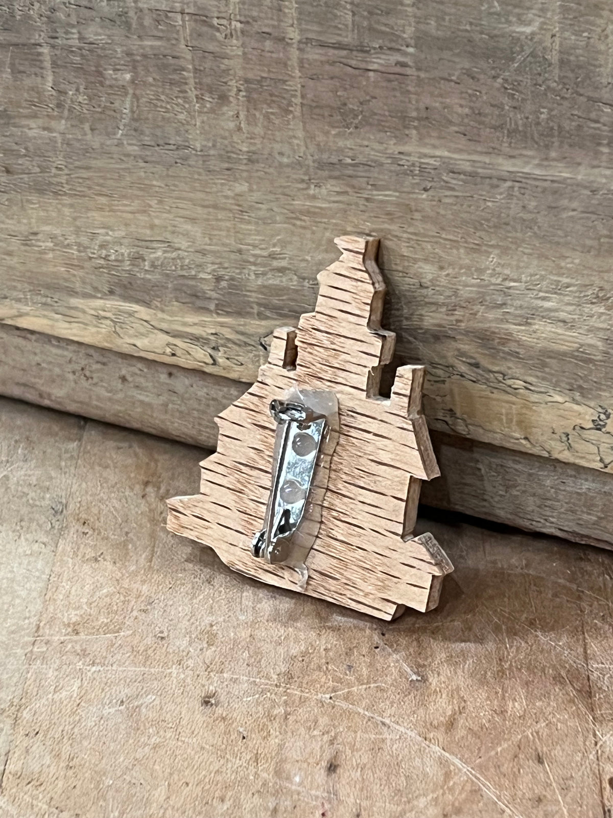 A small wooden Christmas tree-shaped brooch with a silver metal pin back, resting on a rustic wooden surface.
